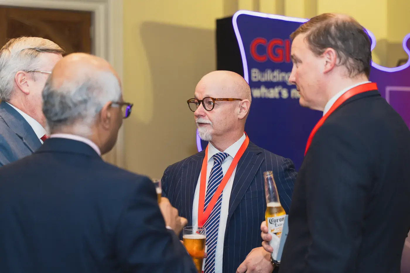 Delegates in suits in conversation, drinks in hand, in front of a lit brand backdrop